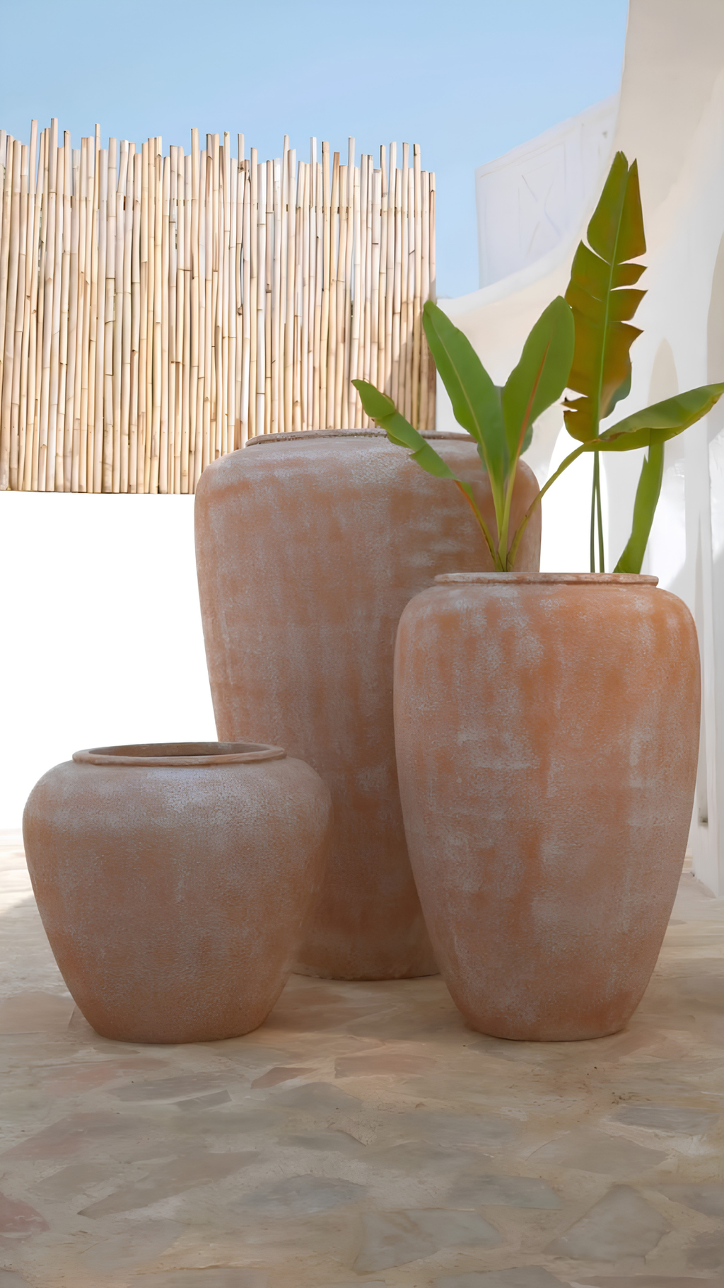 Three terracotta-colored ceramic planters in different sizes arranged on a stone surface, with a green plant in the largest planter. A bamboo screen and white wall are visible in the background.