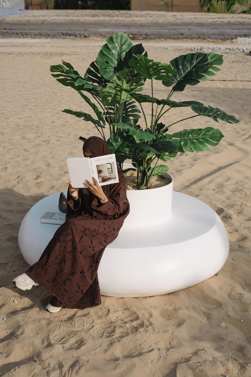 Person sitting on a white  donut shaped planter seater reading a book next to a potted plant on a sandy surface.