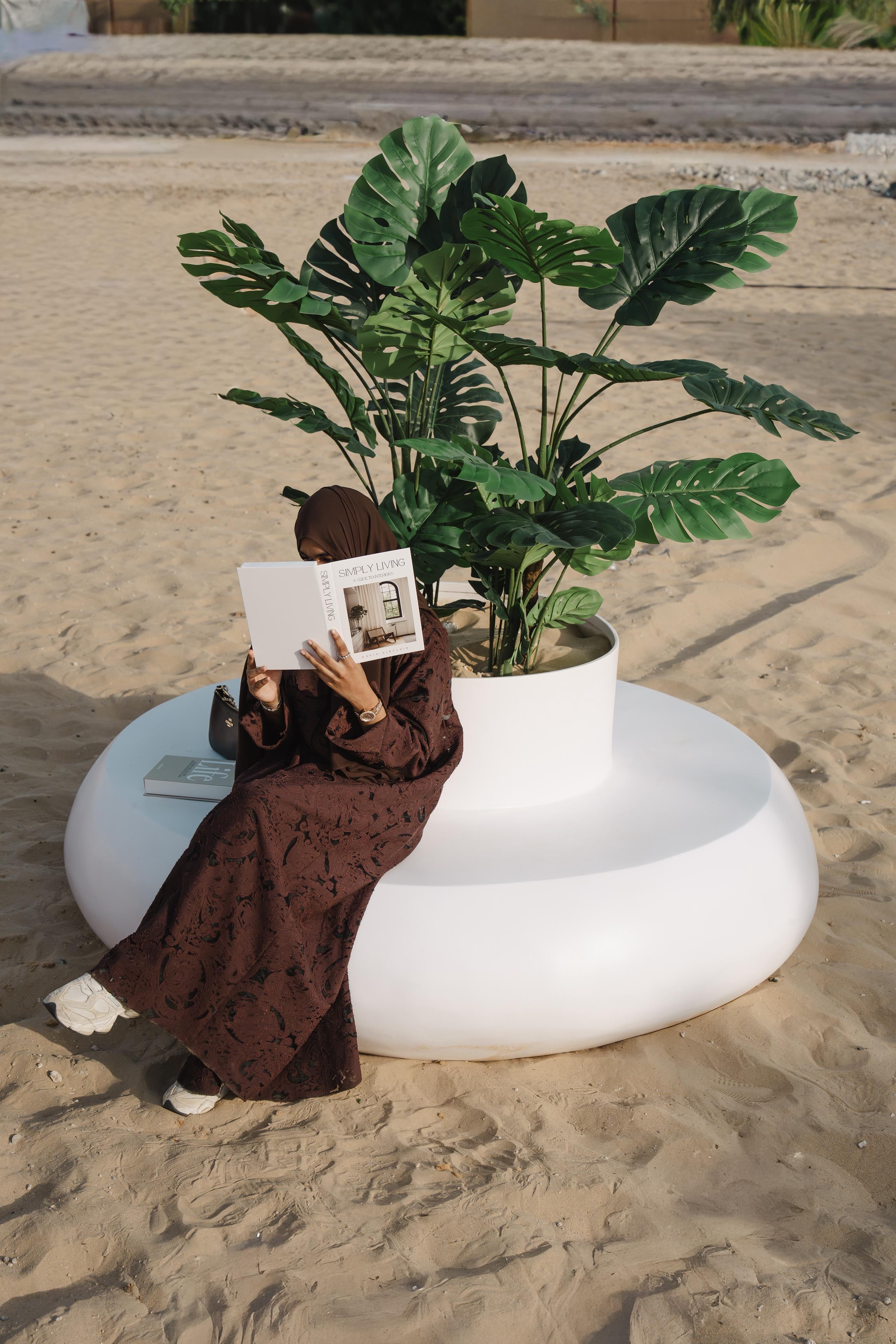 Person sitting on a white  donut shaped planter seater reading a book next to a potted plant on a sandy surface.