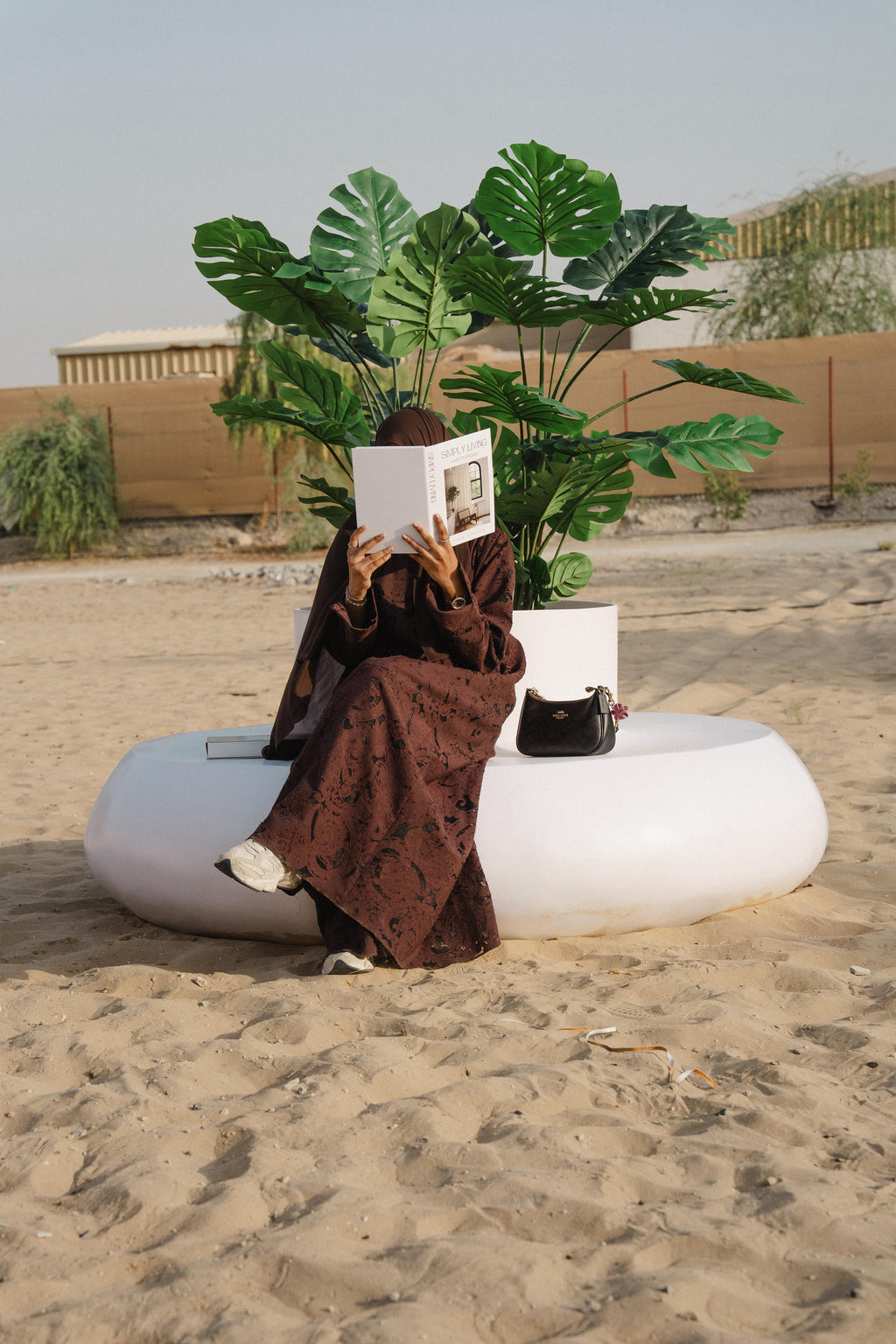 Person sitting on a white donut shaped planter seater in a desert-like setting, reading a book with plants and a building in the background.